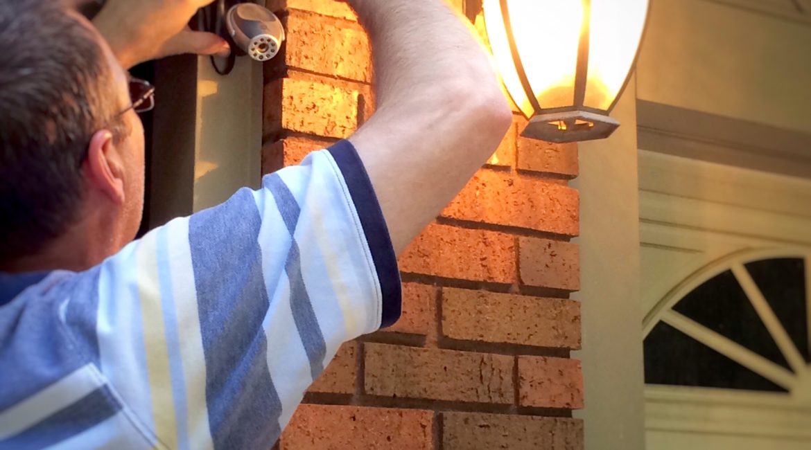 An older white man installs a camera outside of his brick home. A porch light is lit casting a warm glow.